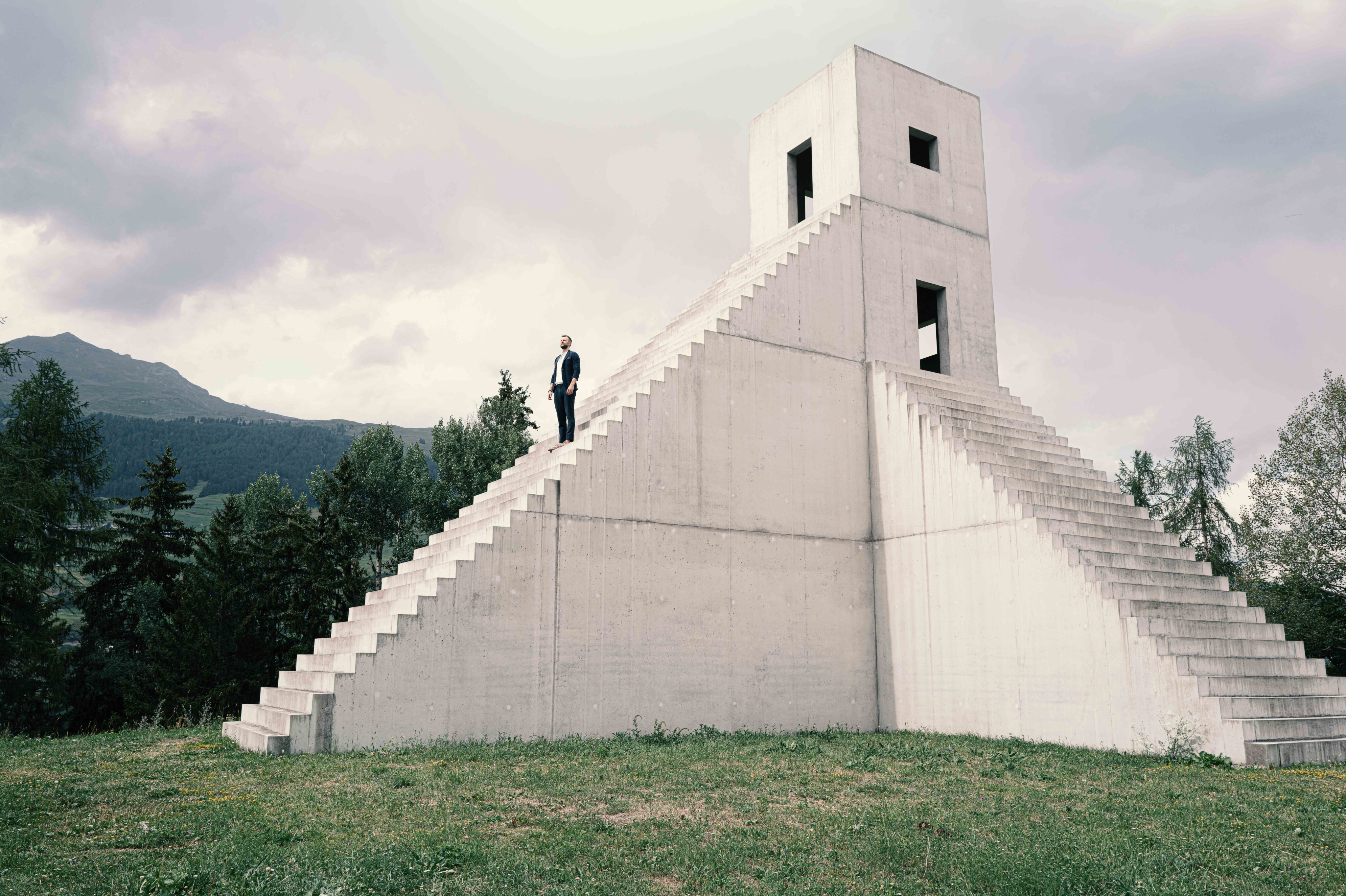 Ein Mann steht auf einer monumentalen, minimalistischen Betontreppe vor einer Bergkulisse, die architektonische Exklusivität und Weite symbolisiert.