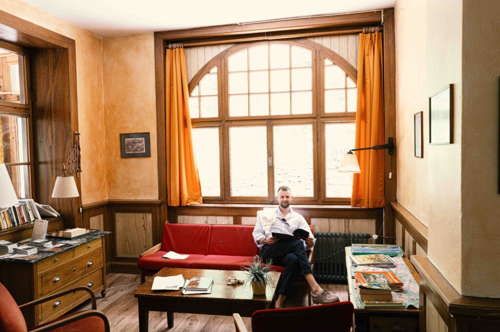 Ein Mann sitzt in einem stilvollen Wohnzimmer mit historischem Rundbogenfenster, rotem Sofa und eleganten Holzvertäfelungen in einer Villa.