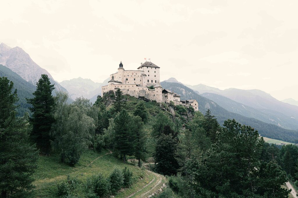 Historische Burg auf einem bewaldeten Bergkamm vor einer imposanten Alpenkulisse, umgeben von grünen Nadelwäldern und einer weiten Berglandschaft.
