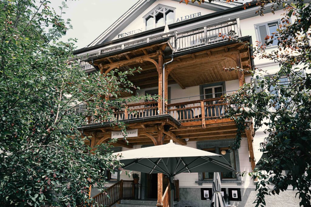 Historische Villa mit aufwendig geschnitzten Holzbalkonen und weißer Fassade, umgeben von üppigem Grün und einem großen Sonnenschirm im Vordergrund.