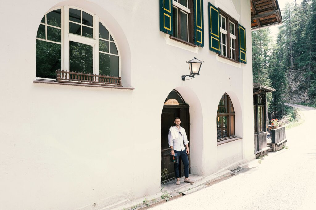 Ein Mann steht vor einer historischen Villa mit weißer Fassade, Rundbogenfenstern und grünen Fensterläden in einer exklusiven Waldumgebung.