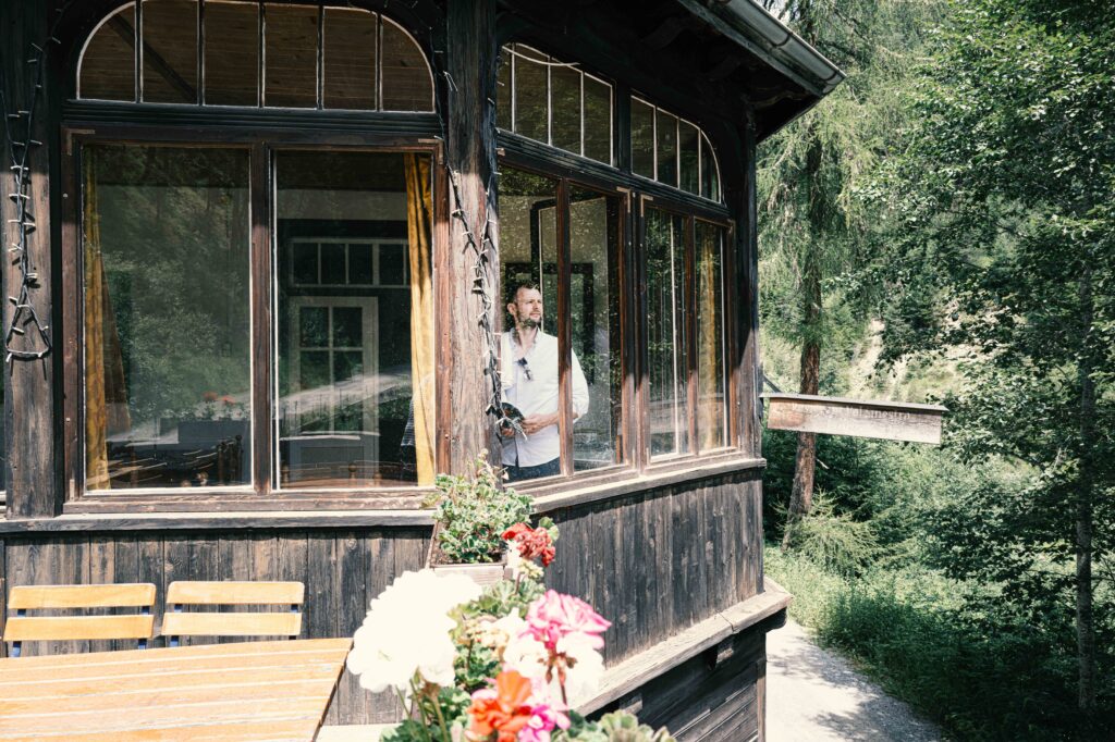 Ein Mann steht in einem historischen Holzhaus mit großen Fenstern und Blick ins Grüne, davor ein Holztisch mit blühenden Geranien im Vordergrund.