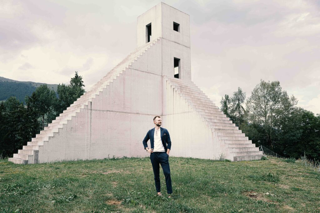 Ein Mann im eleganten Anzug vor einer modernen, pyramidenartigen Betonarchitektur mit markanter Treppenstruktur in einer weitläufigen Landschaft.