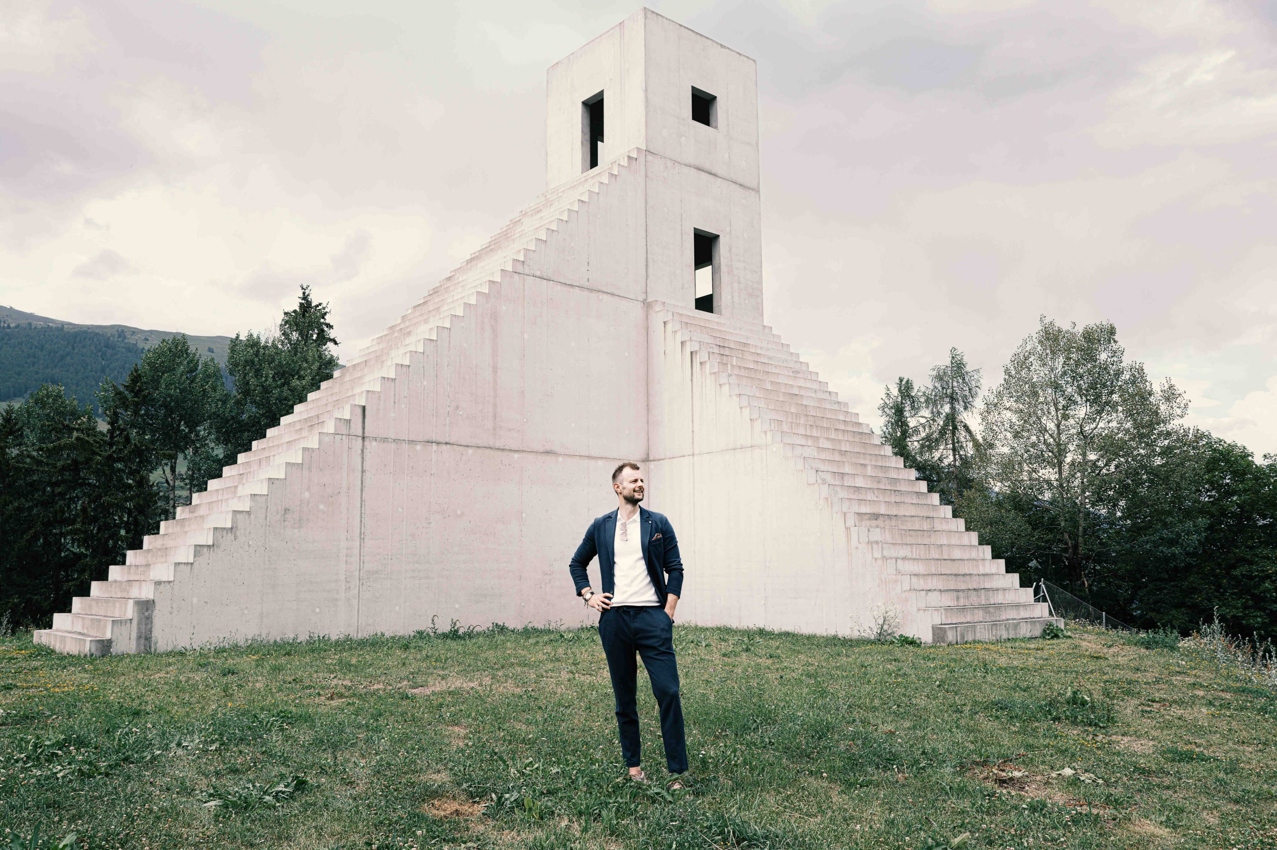 Ein Mann im eleganten Anzug vor einer modernen, pyramidenartigen Betonarchitektur mit markanter Treppenstruktur in einer weitläufigen Landschaft.