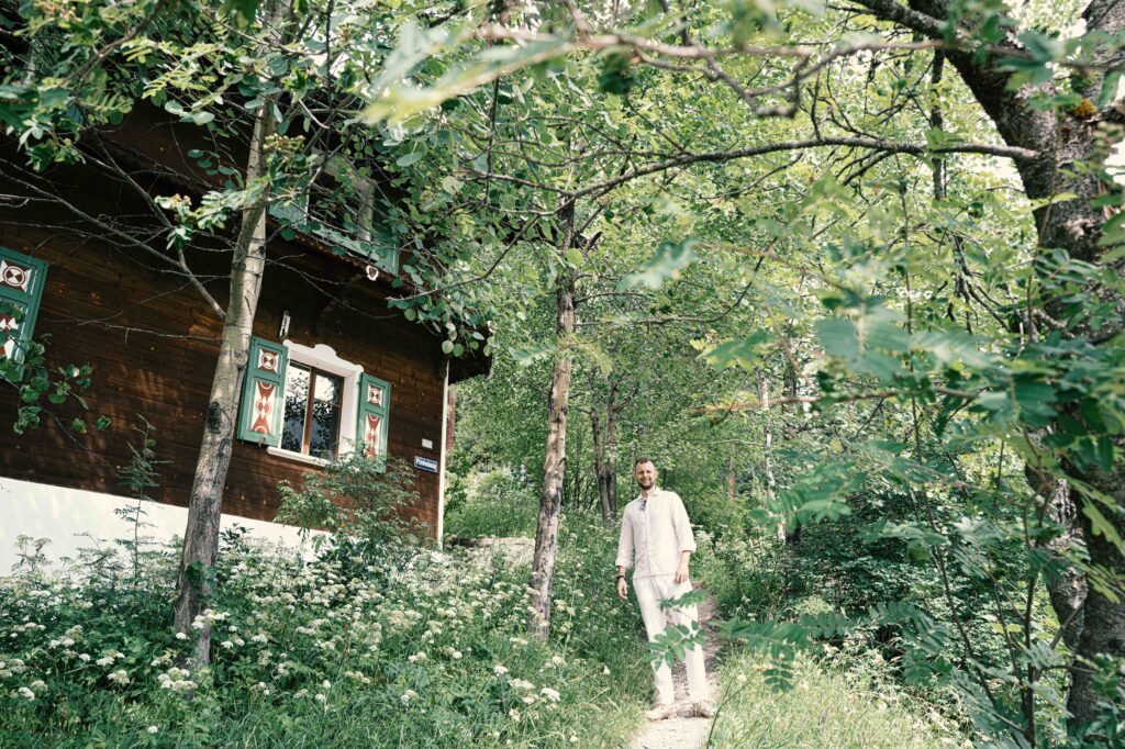 Ein Mann in heller Kleidung steht auf einem Waldweg neben einem rustikalen Holzhaus mit verzierten Fensterläden in einer grünen Gartenanlage.