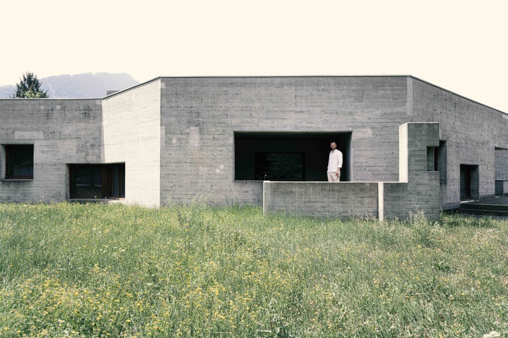 Moderner Sichtbeton-Bau mit markanter Architektur und einer Person auf der Terrasse vor einer blühenden Wiese in einer exklusiven Wohngegend.