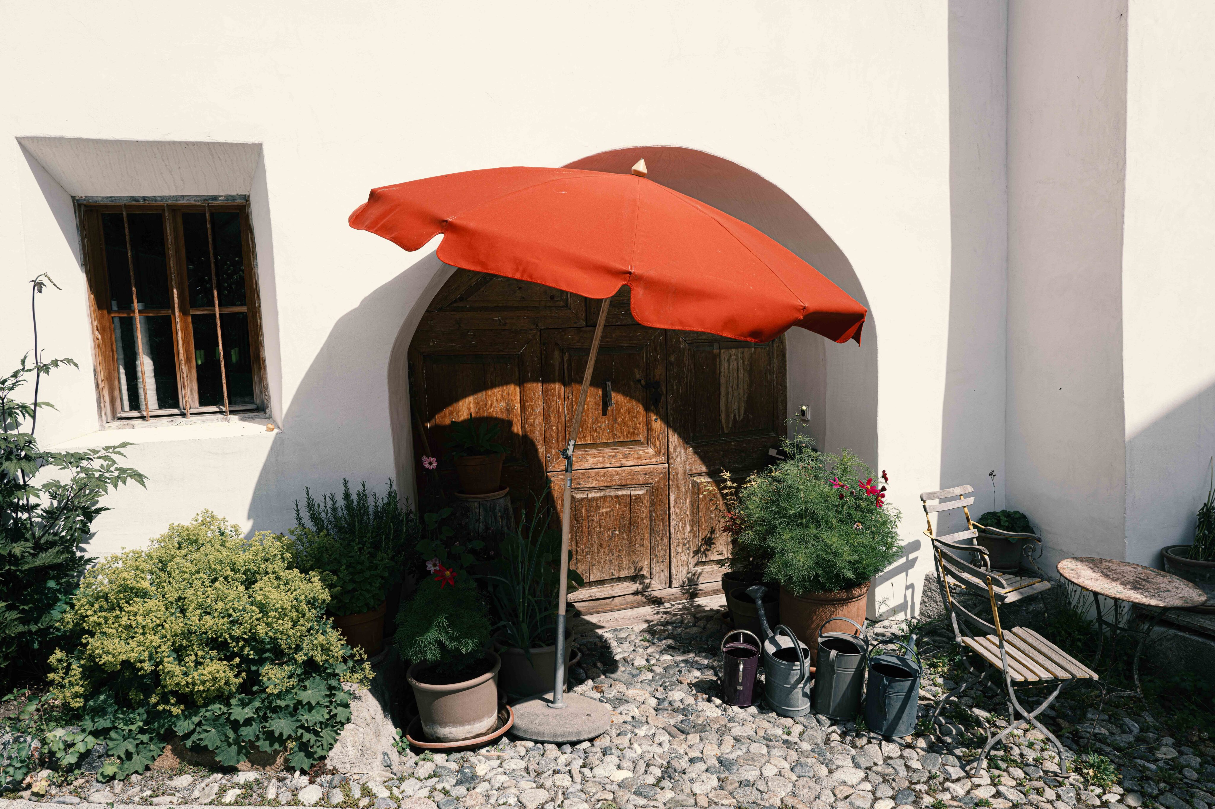 Sonniger Innenhof mit historischem Holztor, Pflanzkübeln und rotem Sonnenschirm.