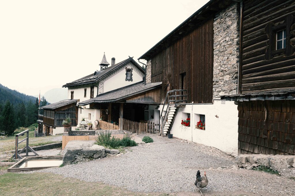 Traditioneller Bauernhof in den Alpen mit rustikaler Holzfassade, weißem Putz, Natursteinelementen und einem kleinen Glockenturm vor Bergkulisse.