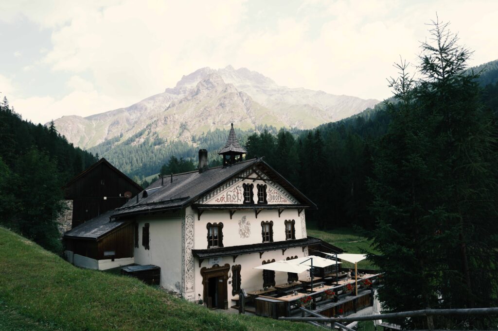 Traditionelles Gasthaus mit kunstvoller Fassadenmalerei vor einer imposanten Bergkulisse in einer ruhigen, grünen Alpenlandschaft.