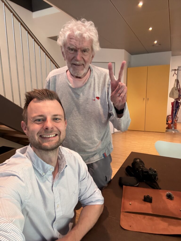 Ein Immobilienmakler und ein älterer Mann lächeln in einem modernen, hellen Wohnraum mit Treppe und gelben Einbauschränken in die Kamera.